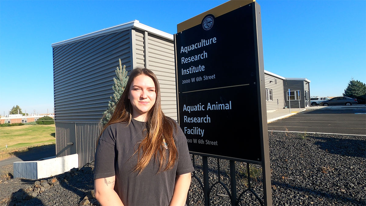 A young woman stands in front of a sign with sun in her eyes.