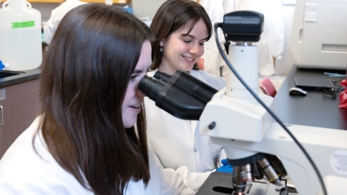 Two female students wearing lab coats peer into microscopes