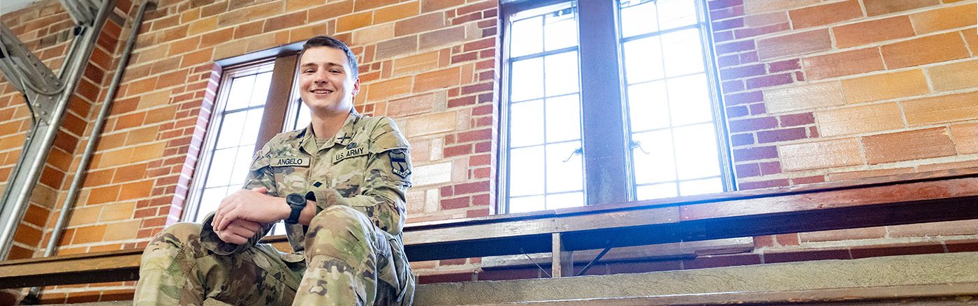 Man wearing military cammies and boots sits on a bench in a brick building.