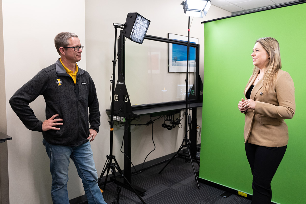 Sean Quallen talks to Mya Groza who stands in front of a green screen in a video production studio filled with recording equipment. 