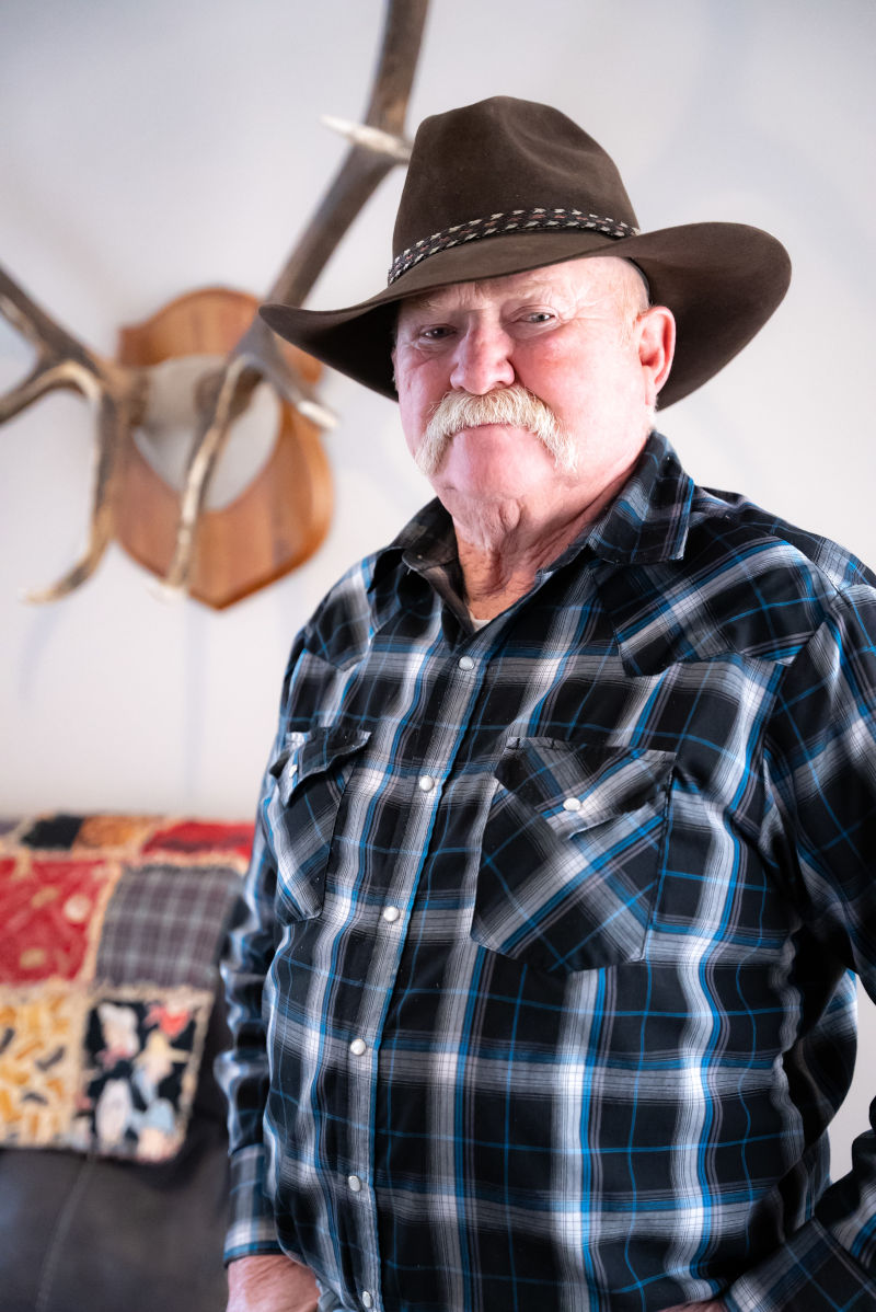 Man in Western hat stands in front of elk antlers.