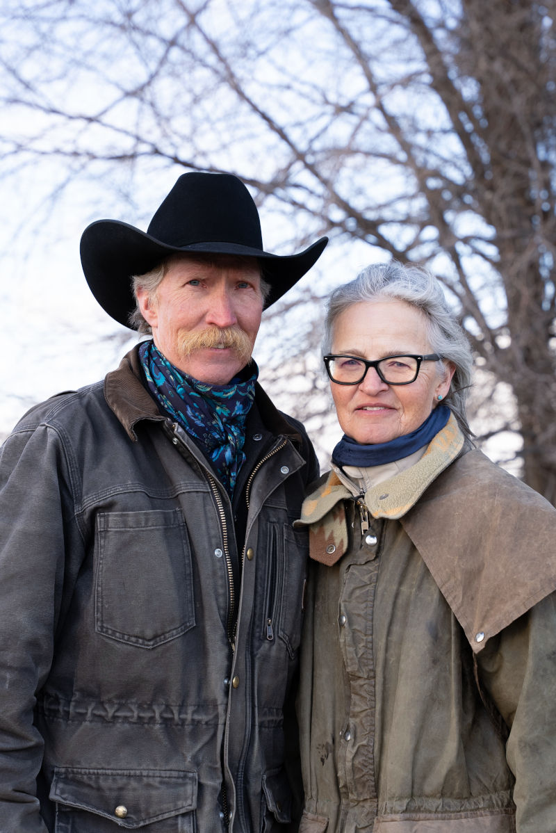 A man with a Western hat and cowboy mustache stands in a field near a woman wearing a duster style ranch coat.