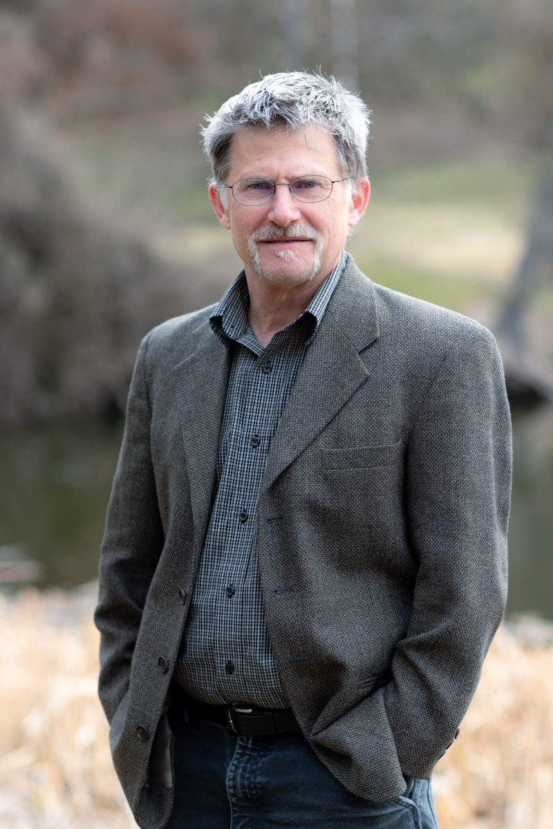 Bespeckled man wearing tweed jacket stands in sage grouse steppe.