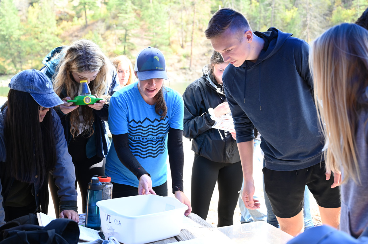 Outreach Specialist Marie Schmidt and high school students stand outdoors and look into a container with lakewater.